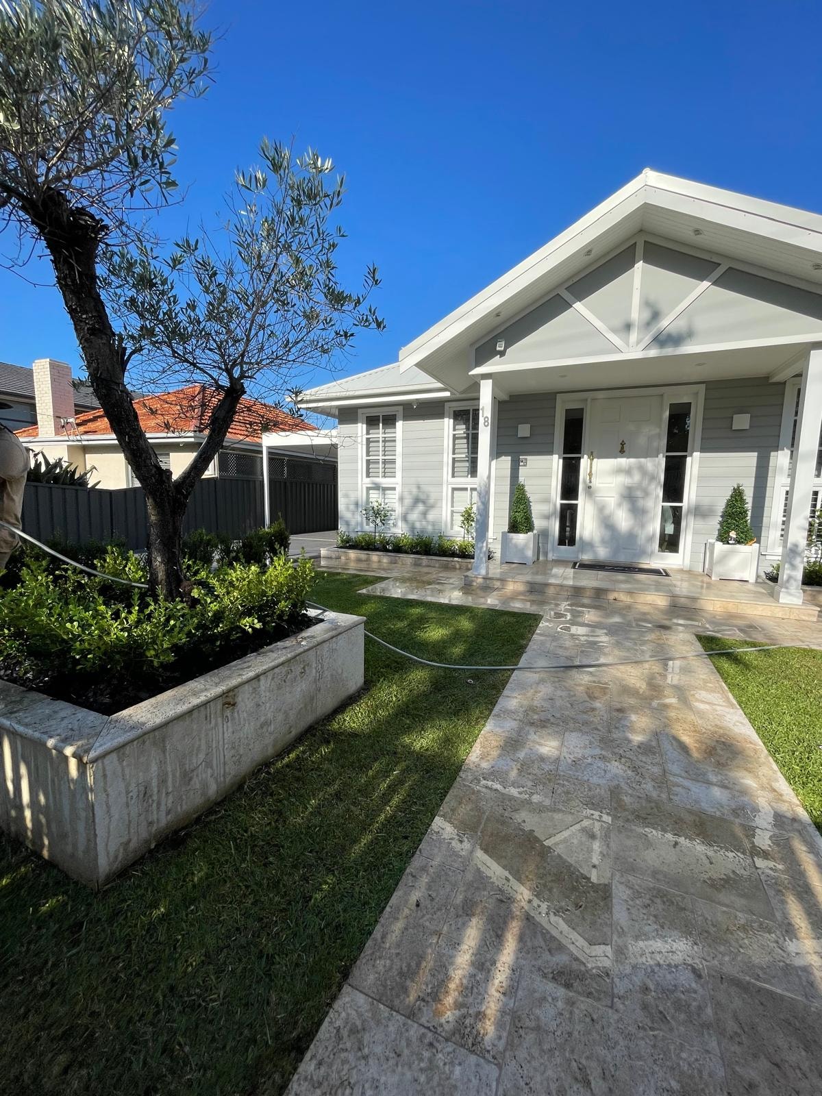 Hamptons-style grey home with travertine path, raised sandstone garden bed, olive tree and lawn