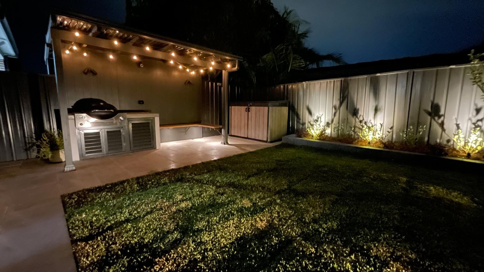 Backyard BBQ patio at night with festoon string lights and garden uplighting