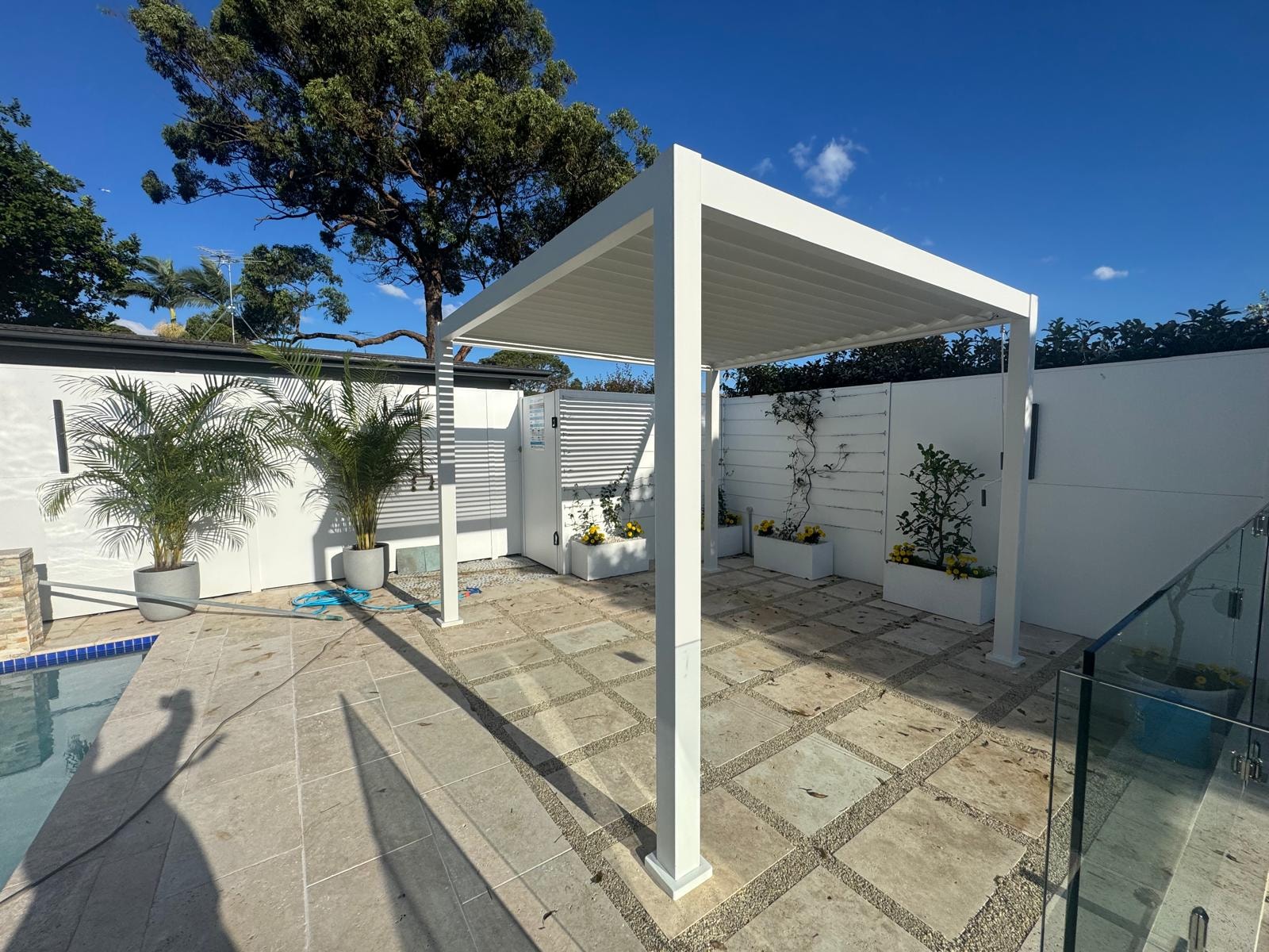 White louvre pergola beside pool with travertine paving and garden planters